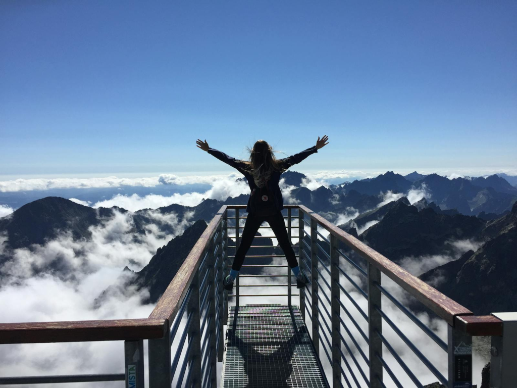 Photo by Nina Uhlikova A woman stands on a viewing platform in Vysoké Tatry, Slovakia, surrounded by clouds and mountains, embracing freedom.