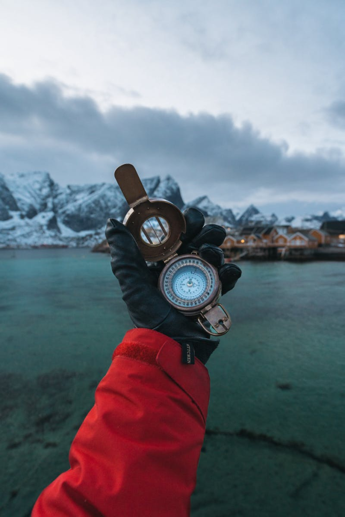 Photo by Simon Migaj A gloved hand holds a compass in Reine, Lofoten Islands, with a stunning fjord backdrop.